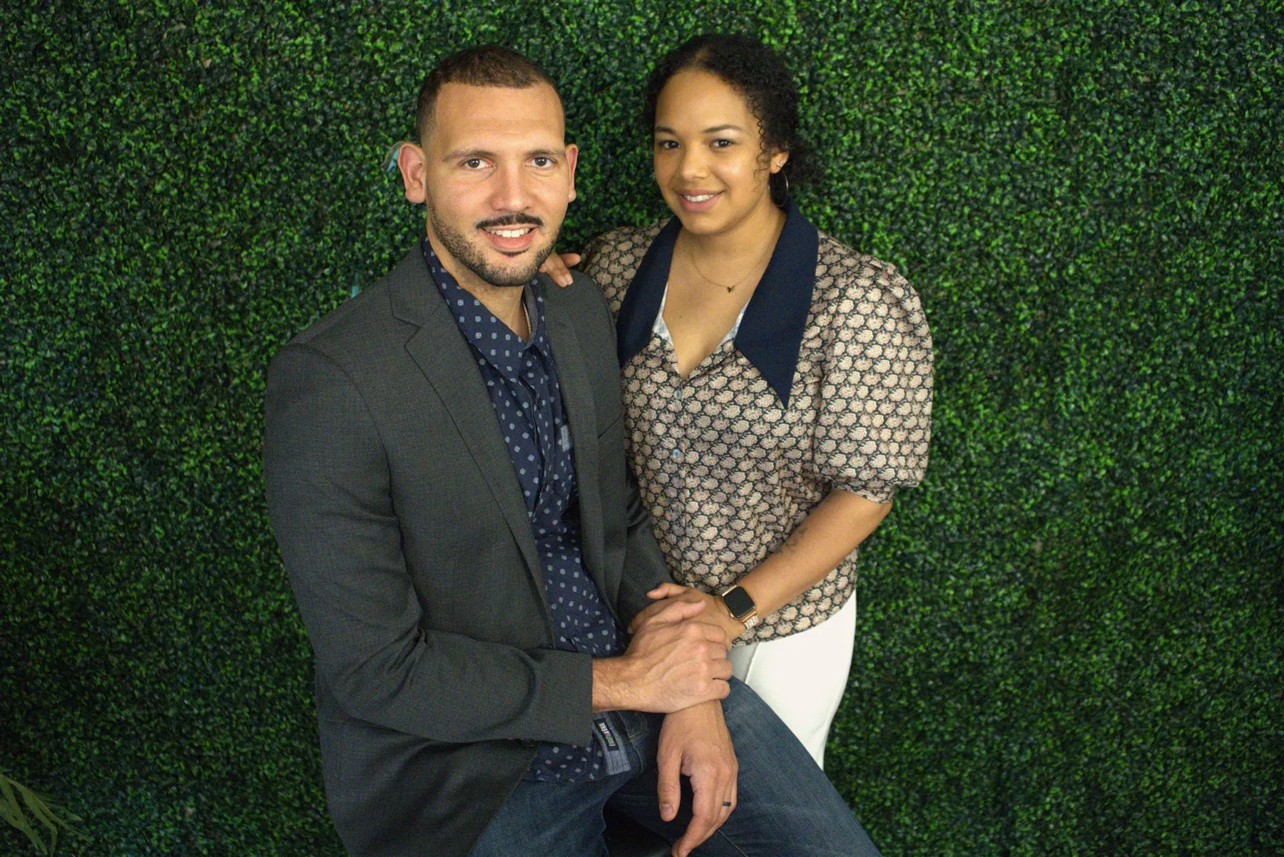Jordan seated and Melina standing beside him in front of a green leafy wall, both smiling